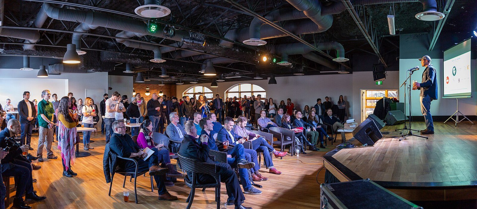 A speaker presents on stage to a large seated and standing audience at a startup pitch night event held in a modern industrial-style venue with exposed ductwork ceilings, wooden floors, and a projected screen visible behind the presenter